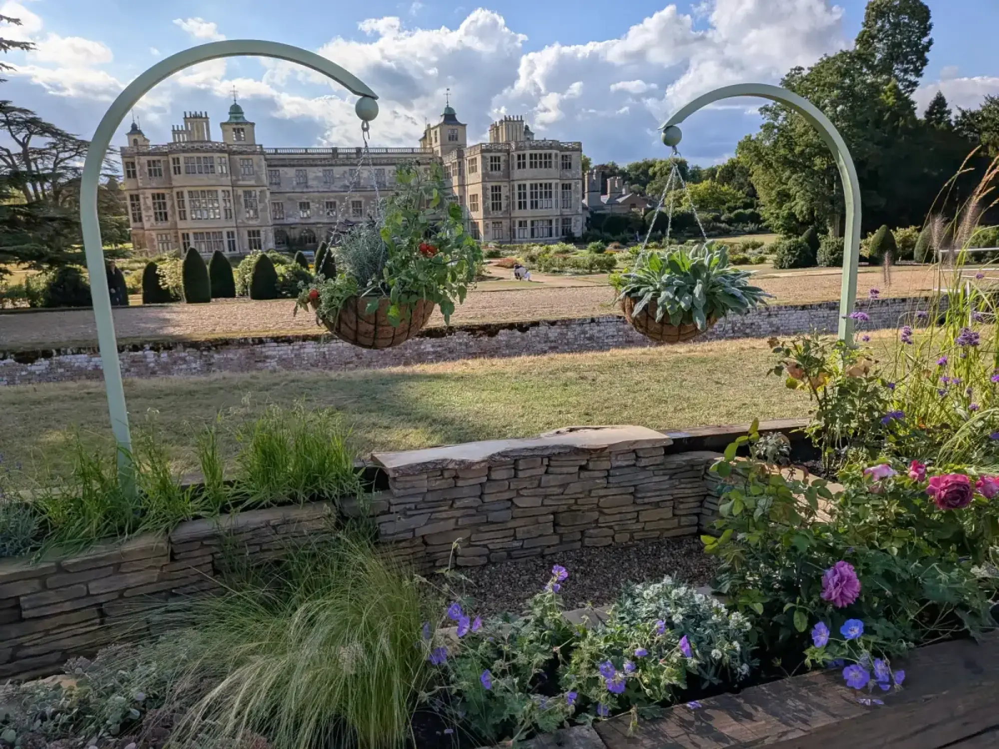 A beautiful border garden with two pale green arches holding hanging baskets, a dry-stacked stone wall, and mixed flowers and grasses.