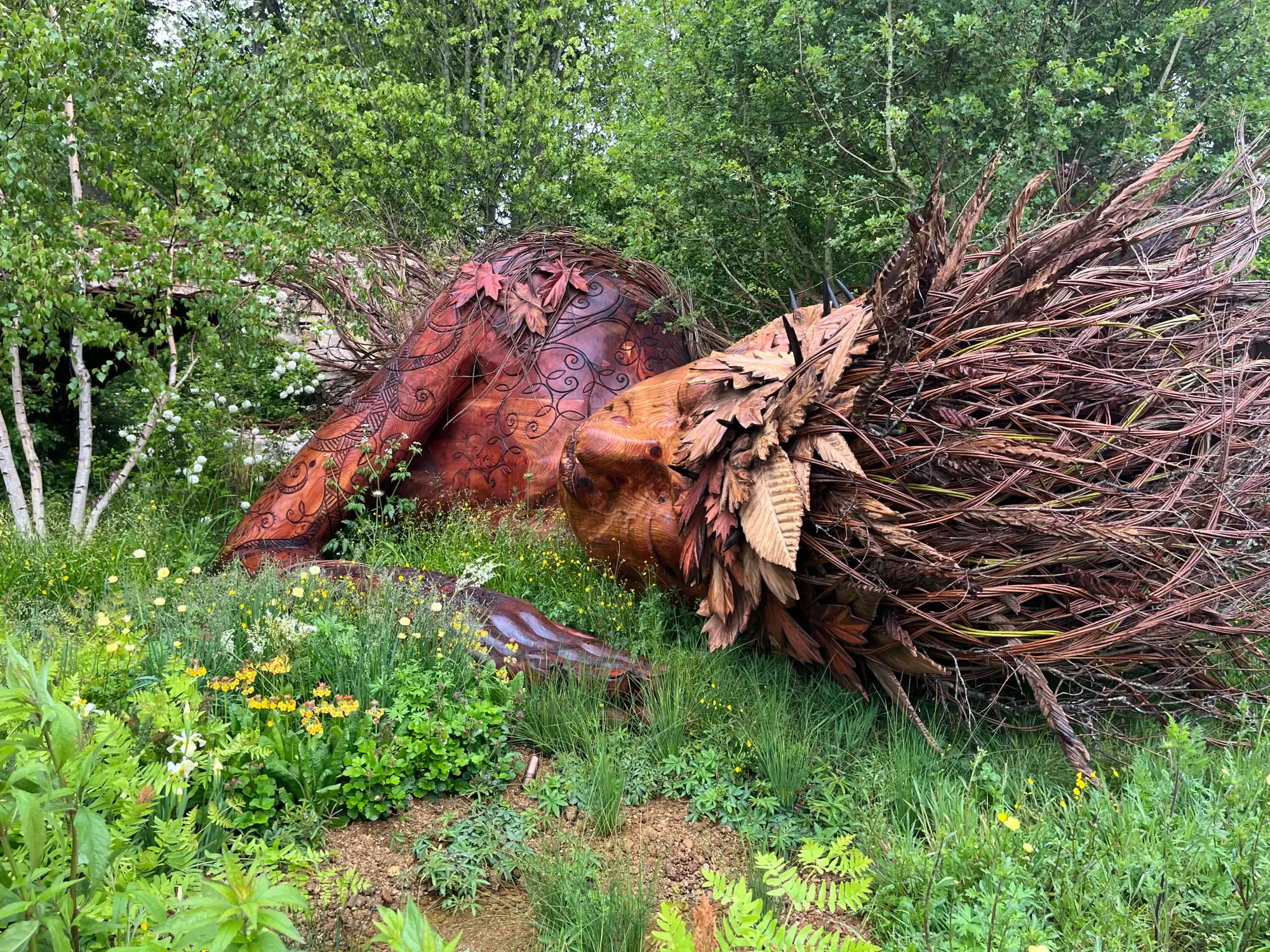 A large carved wooden earth goddess sculpture with branch hair reclines among ferns, grasses, and yellow and white wildflowers.