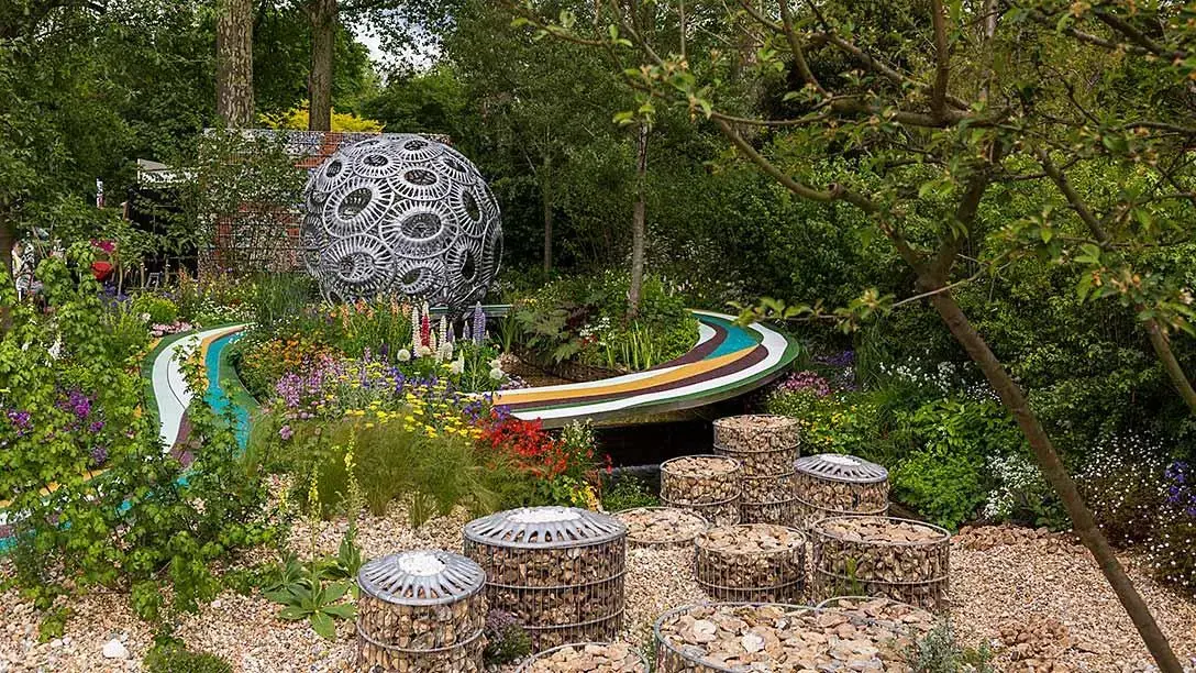 A contemporary show garden with a large spherical metal sculpture, a rainbow-striped path, and stone gabion baskets surrounded by vibrant plants.