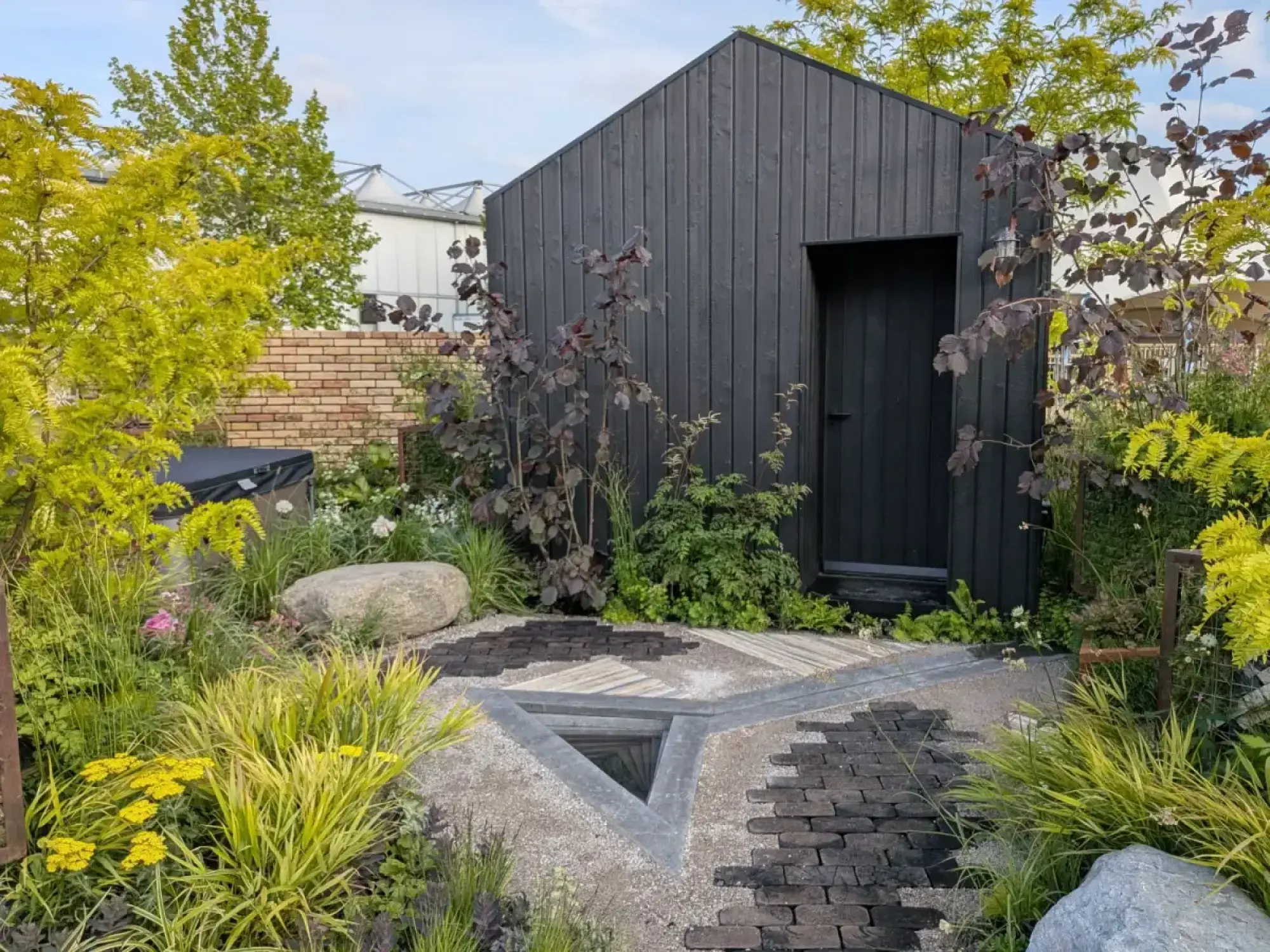 A contemporary show garden features a black timber shed, a triangular water feature, dark brick and gravel paths, and lush planting with lime green, purple, and yellow foliage.