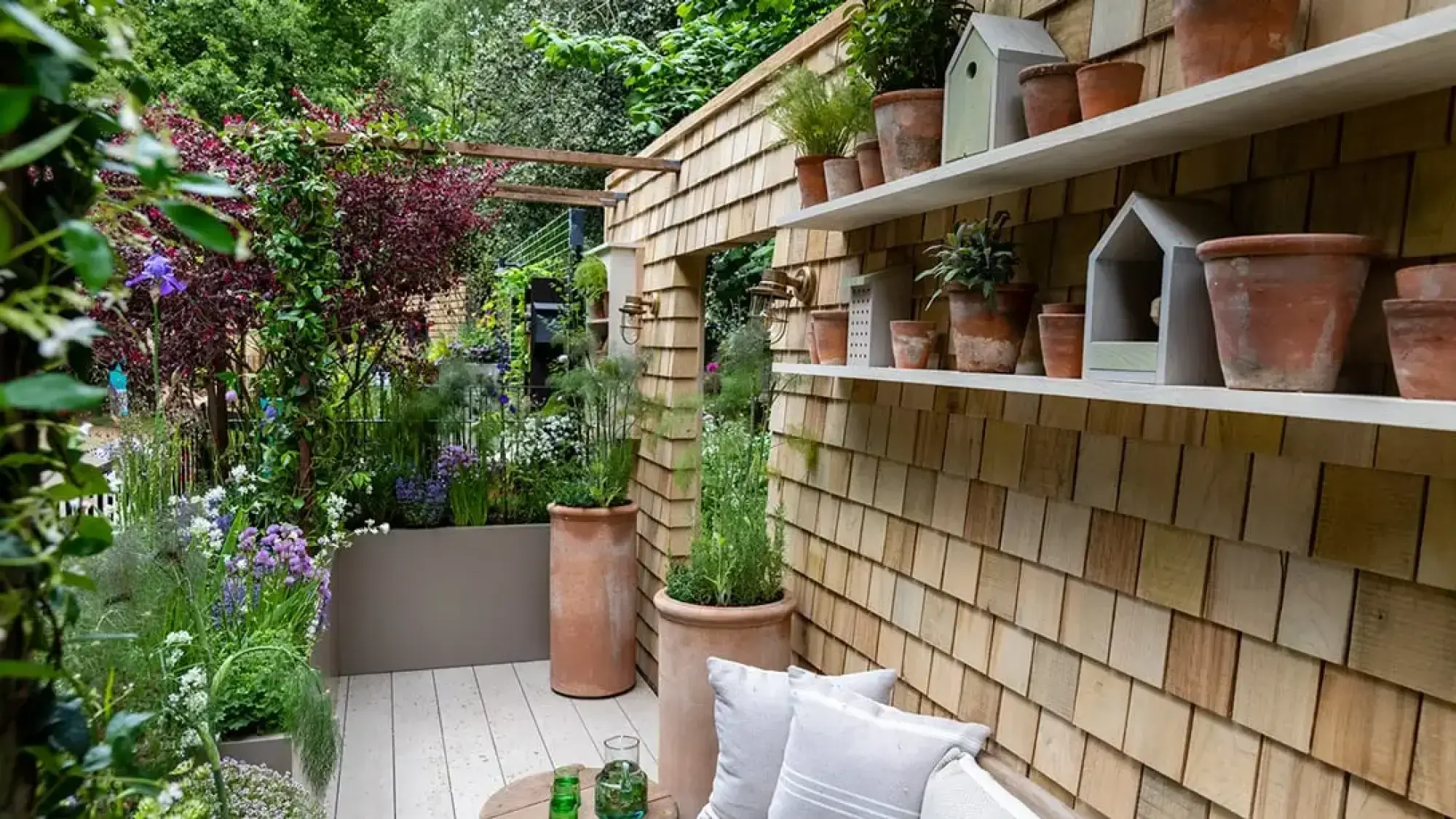 A tranquil balcony garden features a cedar shingle wall with white shelves displaying terracotta pots and decorative birdhouses, lush green, purple, and burgundy planting, and a light wooden deck with outdoor seating.